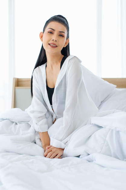 Foto de retrato de uma mulher sexy asiática do milênio em regata de conforto casual e camisa sentada sob cobertor branco grosso e quente posando olhar para a câmera sorrindo depois de acordar no quarto de manhã em casa