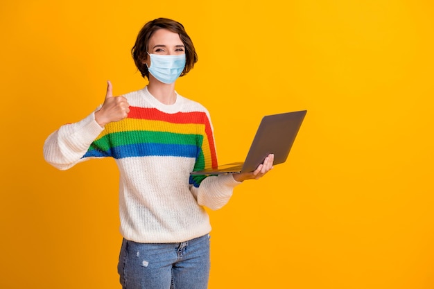 Foto de senhora positiva segurar laptop mostra sinal de aprovação usar máscara médica suéter jeans azul isolado em fundo amarelo