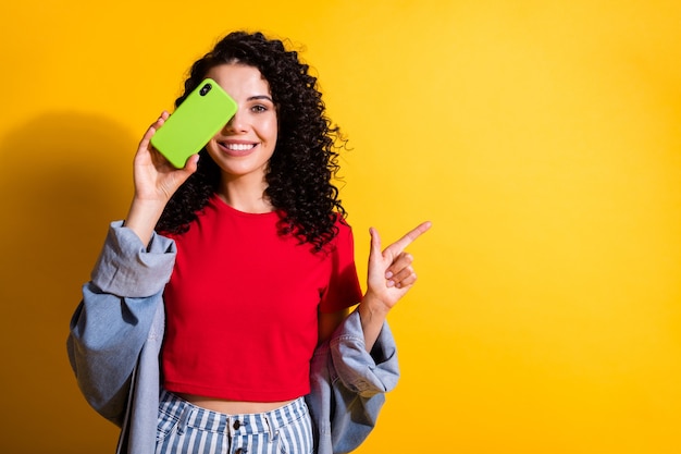 Foto foto de senhora segurar telefone capa olho dedo direto olhar espaço vazio usar camiseta vermelha jaqueta jeans isolada cor amarela de fundo