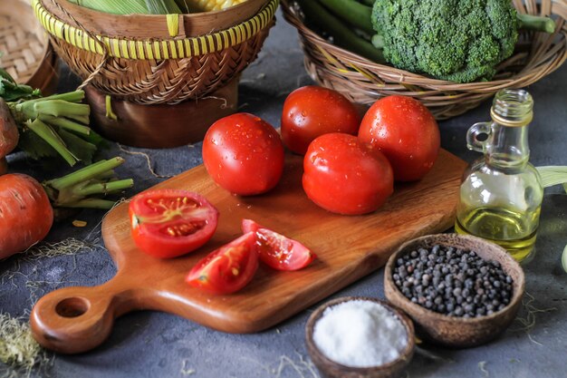 Foto de tomates frescos na tábua, em torno de legumes, cenoura, sal, pimenta do reino, milho, brócolis. Corte os tomates. Colheita de tomates. Mesa de madeira.