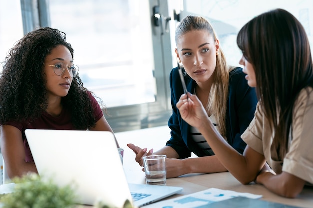 Foto de três empresárias modernas conversando e revisando o trabalho mais recente feito no computador em um espaço de trabalho conjunto