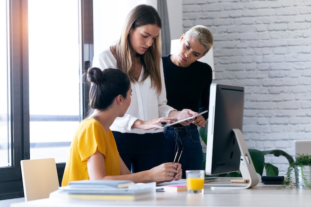 Foto de três empresárias modernas conversando e revisando os últimos trabalhos feitos no tablet digital em um espaço de trabalho conjunto.