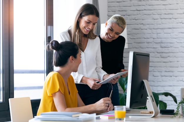 Foto de três empresárias modernas conversando e revisando os últimos trabalhos feitos no tablet digital em um espaço de trabalho conjunto.