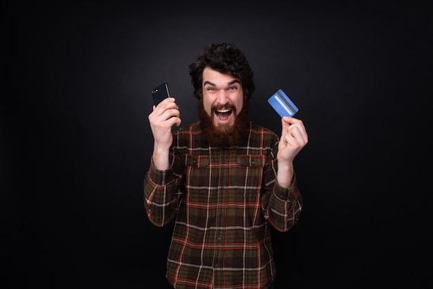 Foto de um barbudo animado com cartão de crédito e smartphone nas mãos, em pé sobre um fundo escuro