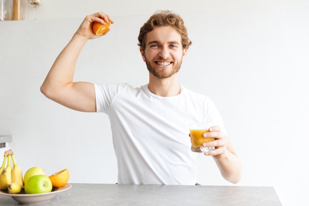Foto de um barbudo jovem feliz na mesa em casa fazer um suco com frutas mostrando o bíceps.