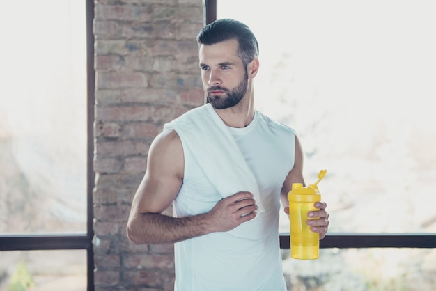 Foto de um belo esportista terminando o treinamento matinal bebendo água doce segurando uma toalha, olhando para as janelas internas da casa de treinamento de roupas esportivas laterais