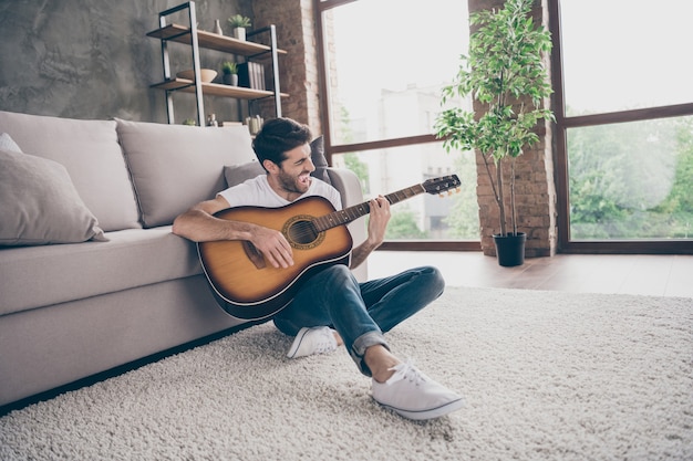 Foto de um cara de raça mista sentado no chão inclinado sofá segurar violão instrumental tocando uma nova música escrita assinatura de som Criativa pessoa talentosa apartamento apartamento sala de estar dentro de casa