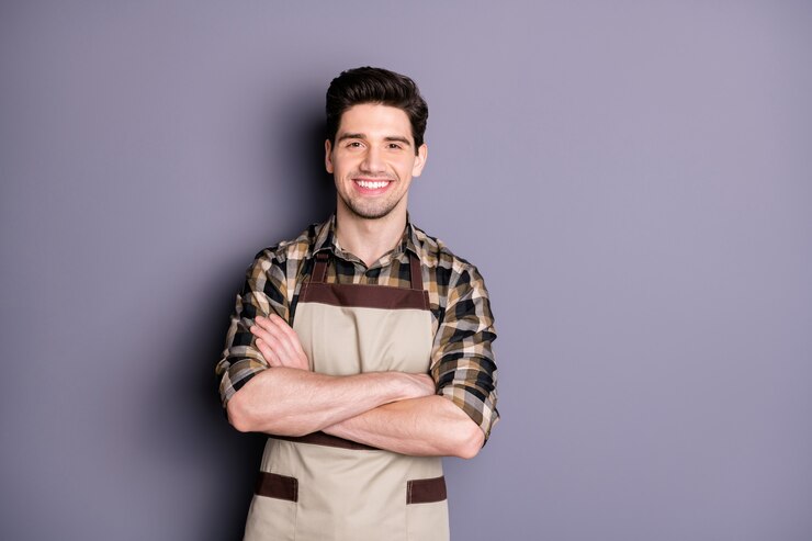 Foto foto de um cara moreno bonito braços cruzados amigável sorrindo positivo bom humor trabalhador profissional usar avental camisa xadrez isolada cor cinza parede