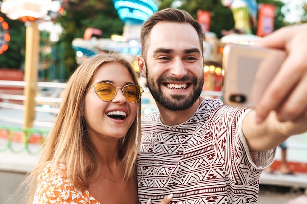 Foto de um casal amoroso sorridente, caminhando ao ar livre no parque de diversões, tire uma selfie pelo telefone celular.