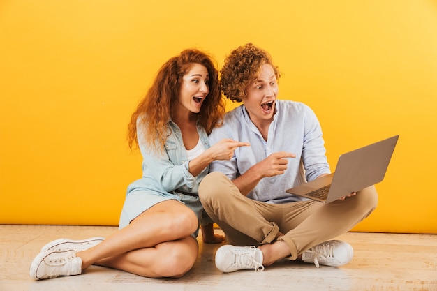 Foto de um casal caucasiano animado e uma mulher sentada no chão e usando um laptop prateado, isolada sobre um fundo amarelo