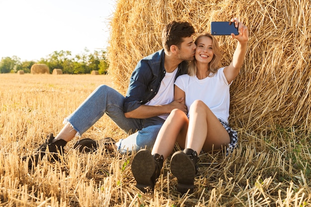 Foto de um casal do campo, um homem e uma mulher tomando selfie enquanto está sentado sob um grande palheiro em campo dourado, durante um dia ensolarado