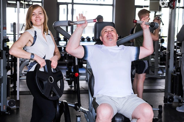 Foto de um casal mais velho no ginásio. Um homem faz um exercício nos braços e ombros.