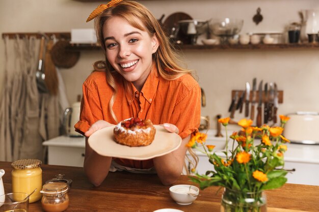 Foto de um chef feliz alegre jovem loira na cozinha segurando o prato com massa doce.