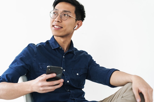 Foto de um empresário chinês alegre usando fones de ouvido, sentado na cadeira e segurando um telefone celular, enquanto trabalhava no escritório, isolado sobre a parede branca