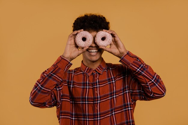 Foto de um fazendeiro africano bonito parece feliz e cobre os olhos por donuts. homem usa camisa xadrez, fundo de cor marrom isolado.