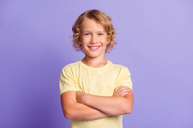 Foto de um garotinho atraente, de mãos cruzadas, olhando para a câmera com um sorriso dentuço isolado sobre um fundo de cor pastel violeta