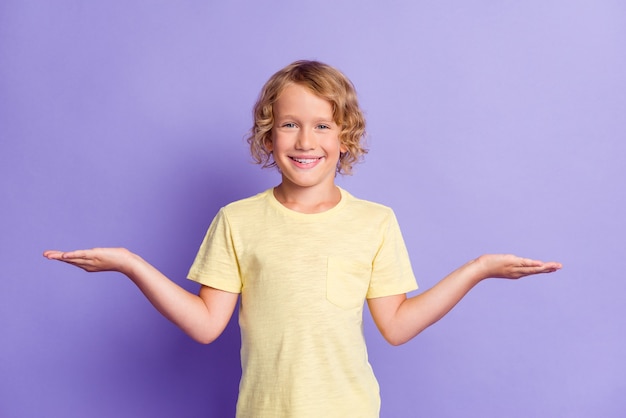 Foto de um garotinho positivo de mãos dadas com o espaço vazio e vestindo uma camiseta amarela isolada sobre um fundo de cor violeta