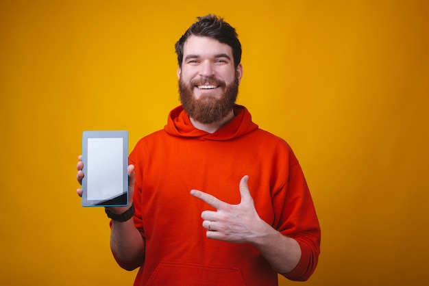 Foto de um hipster barbudo sorridente está mostrando a tela em branco de um tablet no espaço amarelo.