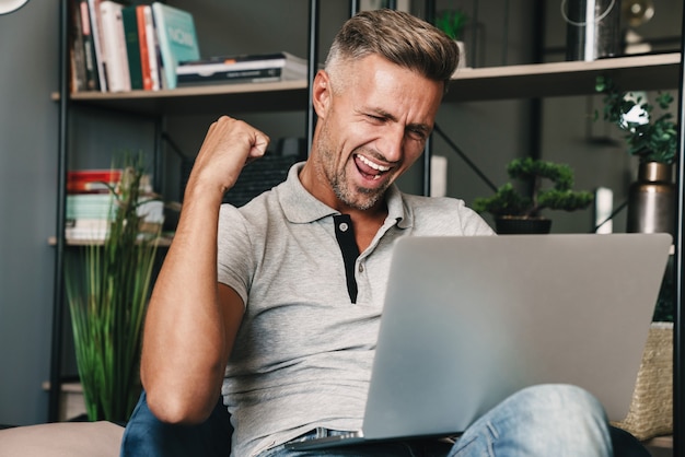 Foto de um homem adulto animado em roupas casuais, sorrindo e comemorando enquanto usa o laptop no apartamento