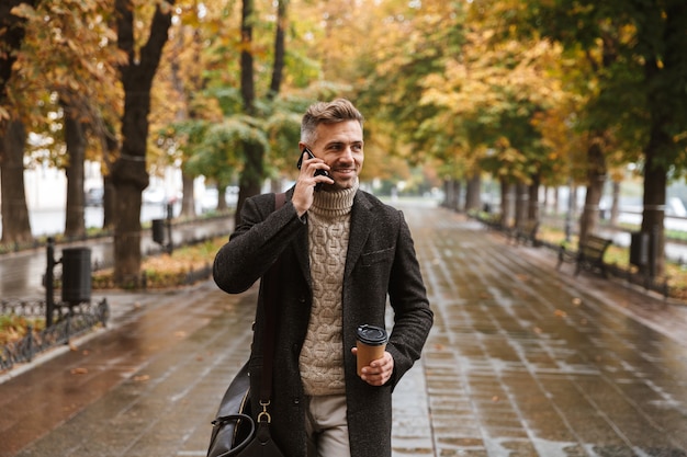 Foto de um homem adulto elegante de 30 anos, vestindo roupas quentes, caminhando ao ar livre pelo parque de outono e usando telefone celular