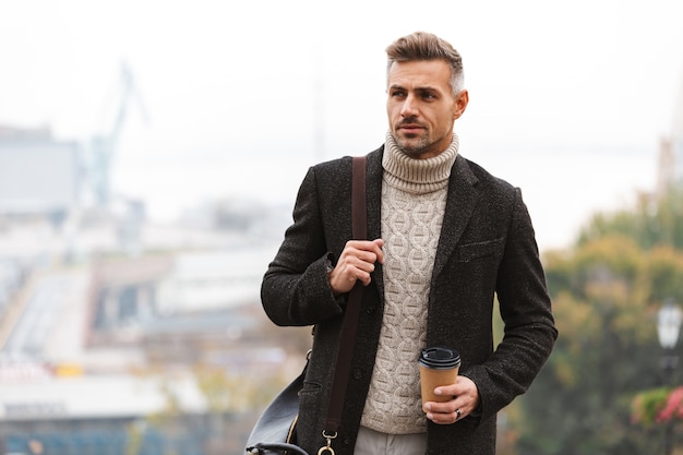 Foto de um homem adulto europeu dos anos 30, vestindo uma jaqueta, segurando um café para viagem, enquanto caminha pela rua da cidade