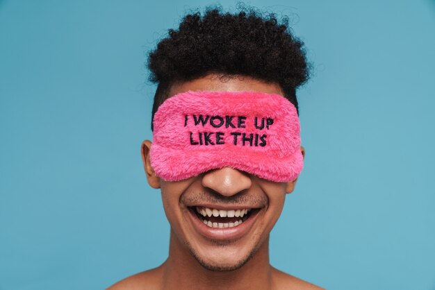 Foto de um homem afro-americano alegre em uma divertida máscara de dormir rindo isolada em um azul