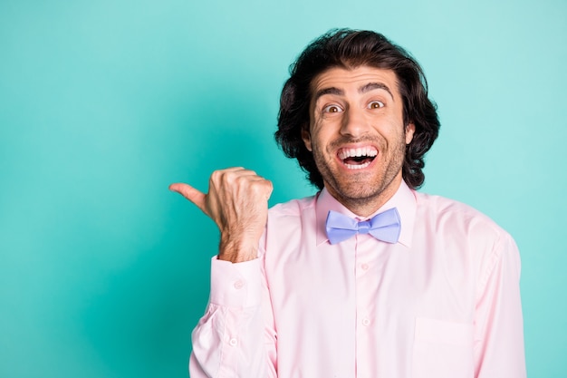 Foto de um homem alegre e cacheado usando uma roupa rosa apontando o dedo polegar para o espaço vazio isolado cor turquesa de fundo