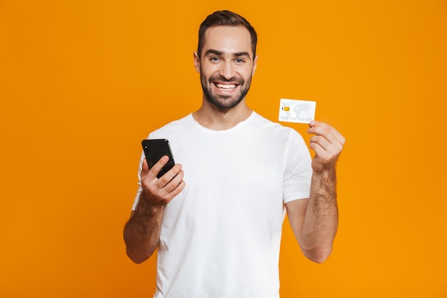 Foto de um homem atraente, com 30 anos, em roupas casuais, segurando um smartphone e um cartão de crédito, isolado