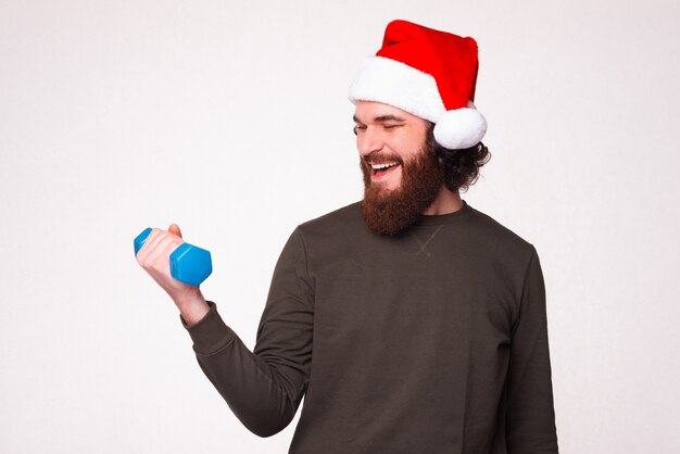 Foto de um homem barbudo feliz com chapéu de Papai Noel e trabalhando com um pequeno haltere azul