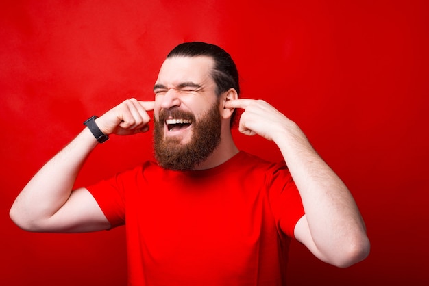 Foto de um homem barbudo frustrado fechando as orelhas com os dedos na parede vermelha