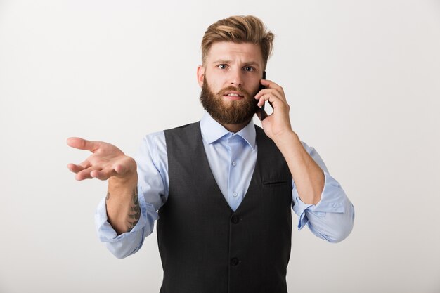 Foto de um homem barbudo jovem sério confuso em pé isolado sobre o fundo da parede branca, falando por telefone celular.