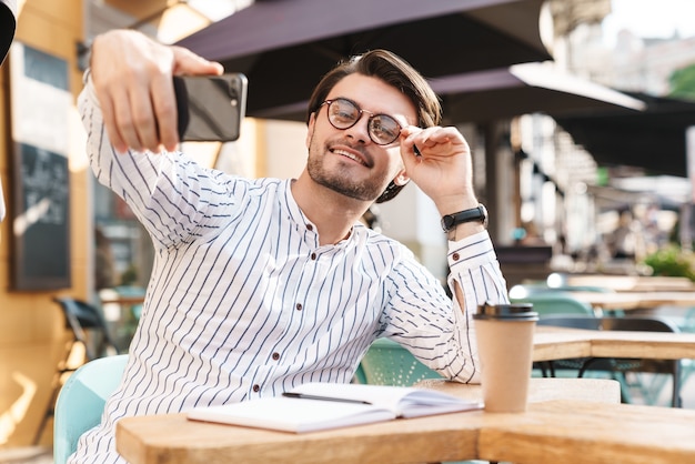 Foto foto de um homem bonito sorridente usando óculos, bebendo café e tirando uma foto de selfie no celular enquanto trabalhava em um café ao ar livre