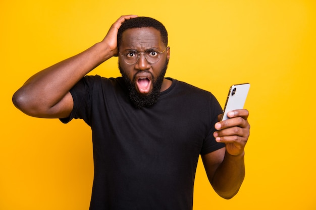 Foto de um homem casual segurando a cabeça depois de ver notícias falsas se espalhando enquanto navega pelo telefone em uma parede de cor amarela vívida isolada de camiseta