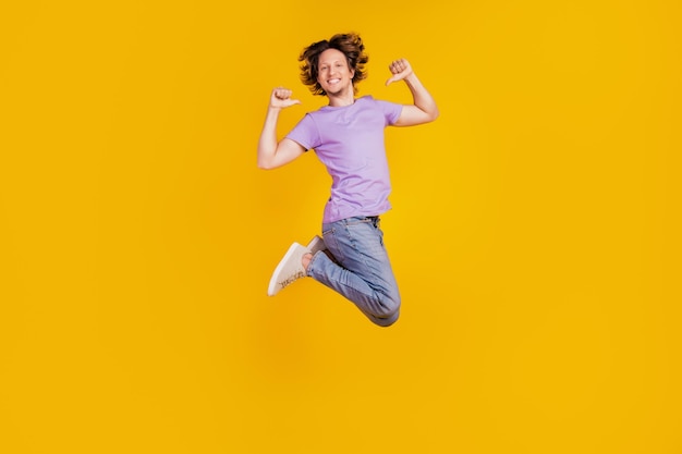 Foto de um homem confiante positivo pulando direto com os dedos do polegar usando roupas jeans casuais em fundo amarelo