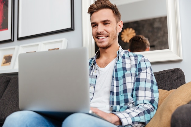 Foto de um homem de cerdas feliz, vestido com uma camisa e estampado em uma gaiola, sentado no sofá em casa e usando o laptop