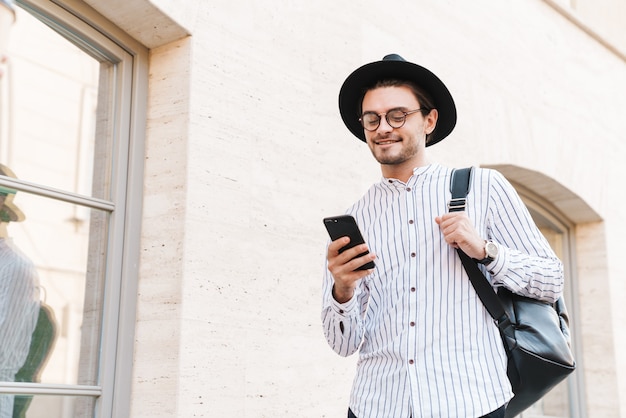 Foto de um homem engraçado bonito usando óculos e chapéu preto, digitando no celular e sorrindo enquanto caminha pela rua da cidade