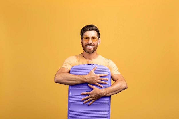 Foto de um homem europeu de meia idade positivo abraçando sua nova mala de bagagem para viagem em pé