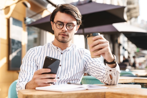 Foto de um homem focado com a barba por fazer usando óculos, digitando no celular e bebendo café enquanto trabalhava em um café ao ar livre