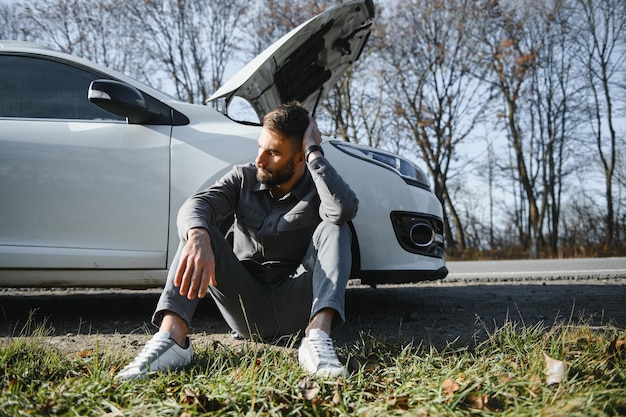 Foto de um homem frustrado sentado ao lado de um carro quebrado com o capô aberto