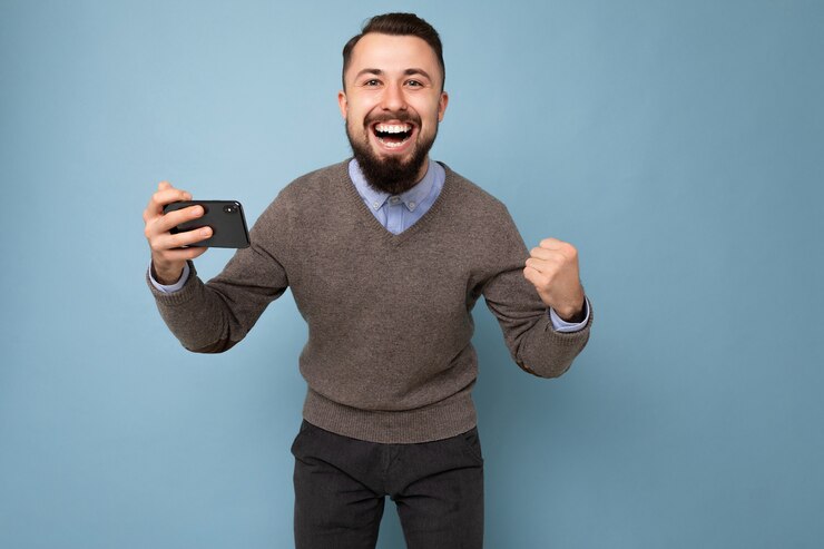 Foto foto de um homem jovem e feliz com barba por fazer, uma jovem morena com barba, vestindo um suéter cinza e uma camisa azul todos os dias, isolada na parede do fundo segurando um smartphone, olhando para a câmera, se divertindo e gritando, sim