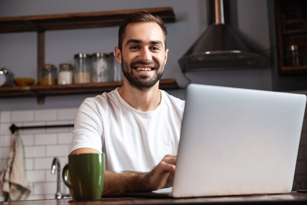 Foto de um homem maduro de 30 anos vestindo uma camiseta casual usando um laptop prateado enquanto está sentado à mesa em um apartamento moderno