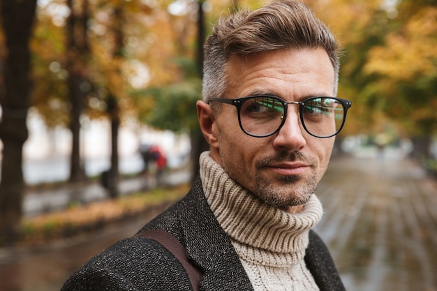 Foto de um homem masculino de 30 anos usando óculos, caminhando ao ar livre pelo parque de outono
