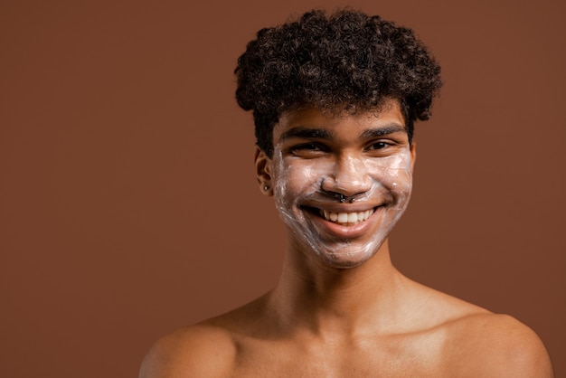 Foto de um homem negro atraente com máscara de creme ou nutrição no rosto, sorri muito bem. Torso nu, fundo de cor marrom isolado.