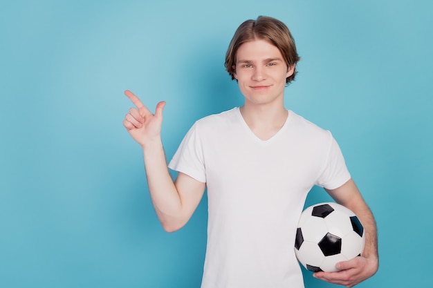 Foto de um homem positivo apontar o espaço vazio com uma bola de futebol isolada no fundo azul