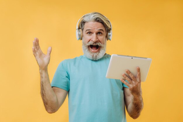 Foto de um homem sênior grisalho expressivo em fones de ouvido com um tablet nas mãos isolado em um fundo amarelo