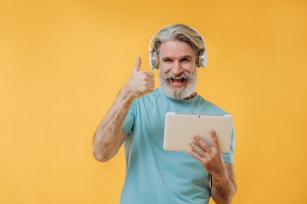 Foto de um homem sênior grisalho expressivo em fones de ouvido com um tablet nas mãos isolado em um fundo amarelo