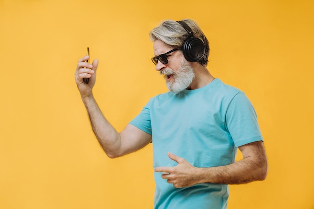 Foto de um homem sênior grisalho expressivo em fones de ouvido com um telefone nas mãos isolado em um fundo amarelo