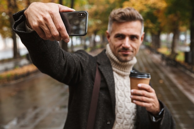 Foto de um homem sorridente de 30 anos, vestindo roupas quentes, caminhando ao ar livre pelo parque outono e tirando uma selfie no celular