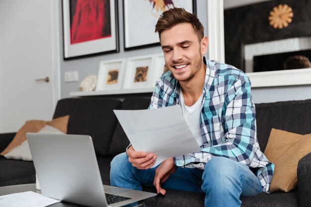 Foto de um jovem alegre e eriçado, vestido com uma camisa e uma gaiola estampada, sentado no sofá em casa e usando o laptop