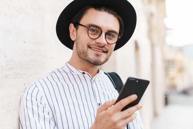 Foto de um jovem alegre usando óculos e chapéu preto digitando no celular e sorrindo enquanto se inclina na parede na rua da cidade